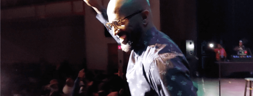 Man on stage, smiling, wearing sunglasses, performing.